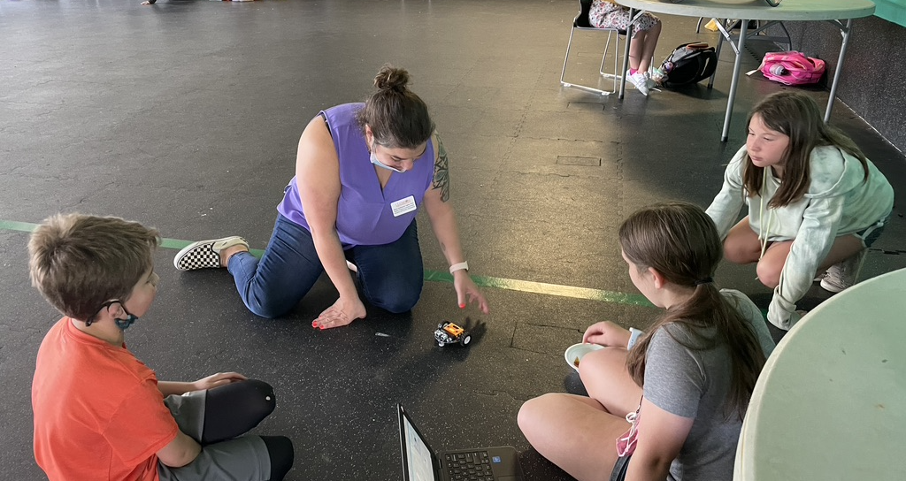A group of kids sitting on the floor with an adult programming a robot using a laptop
